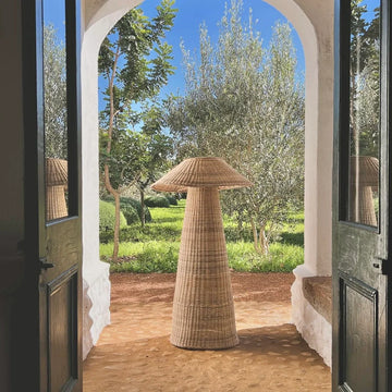 Intaba Cane Woven Floor Lamp standing in an archway overlooking a lush garden, showcasing its elegant design and natural materials.